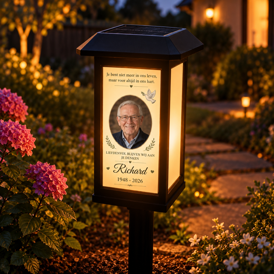 MemoraGarden solar gedenklamp in de tuin bij avondlicht: zwarte lantaarn met een verlichte gedenkplaat met foto, naam en jaartallen tussen de bloemen